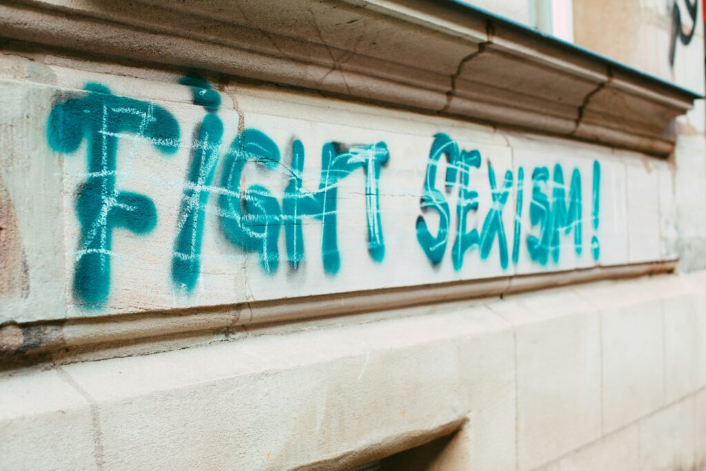 un graffiti "Fight sexism" bleu ciel sur un mur blanc, probablement au dessus d'une porte ou d'un passage